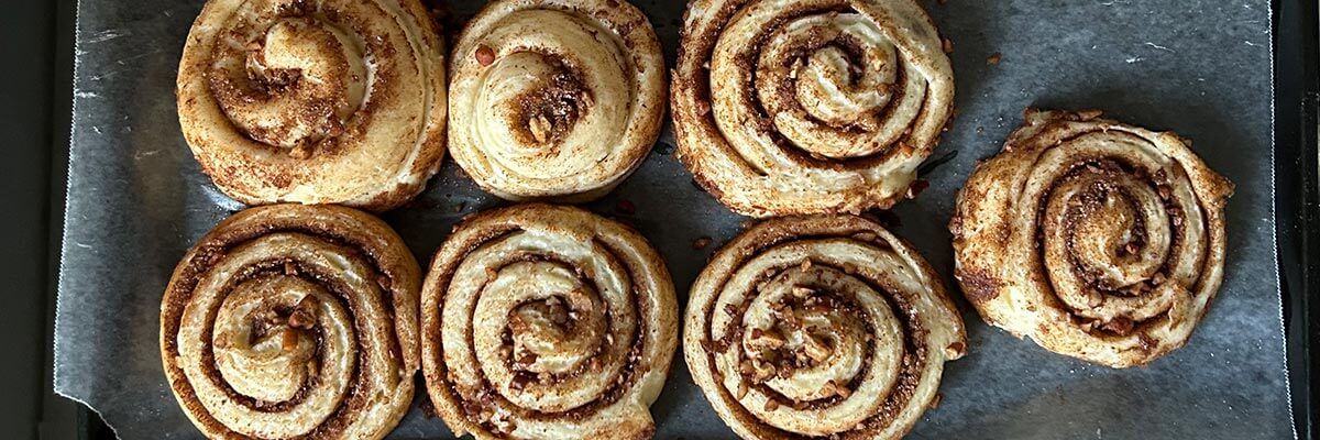 Seven swirled cinnamon buns are photographed from above. They are on a baking pan lined with parchment paper.