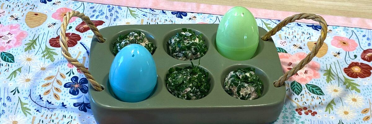 A green plastic tray with a rope handle on each side sits on a floral table runner. The tray has six round compartments in two rows of three. There is a blue plastic egg in the bottom left compartment and a green plastic egg in the top right compartment. The other compartments hold small round appetizers coated in dark green parsley.