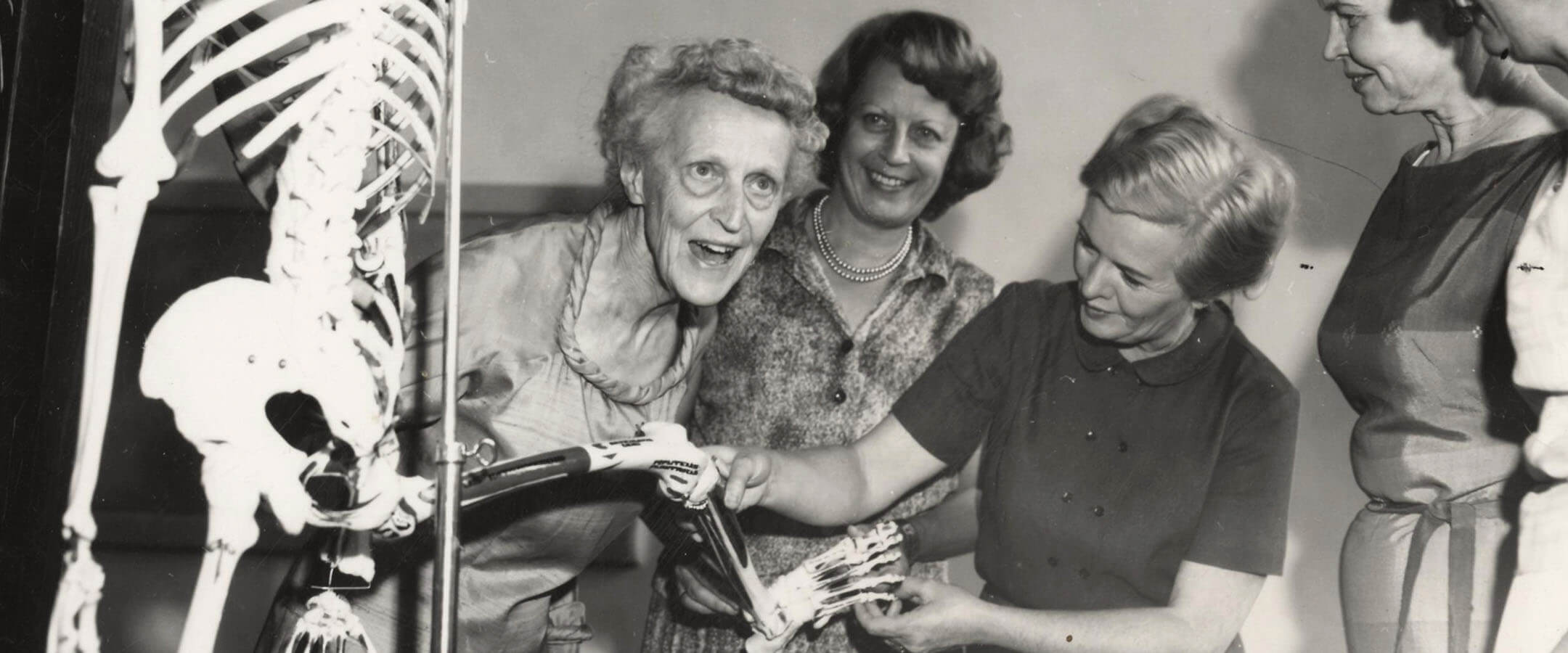 A black and white image of three women and a teaching skeleton. On the left, the torso and pelvis of a teaching skeleton next to the pole on which it is displayed. To the right of the skeleton, an old woman with short, curly hair leans toward the right smiling at someone off camera. To her right, a younger woman with short hair and a floral blouse smiles at her. To her right, another woman in a dark, short-sleeve, button-up blouse holds and examines a leg of the teaching skeleton.