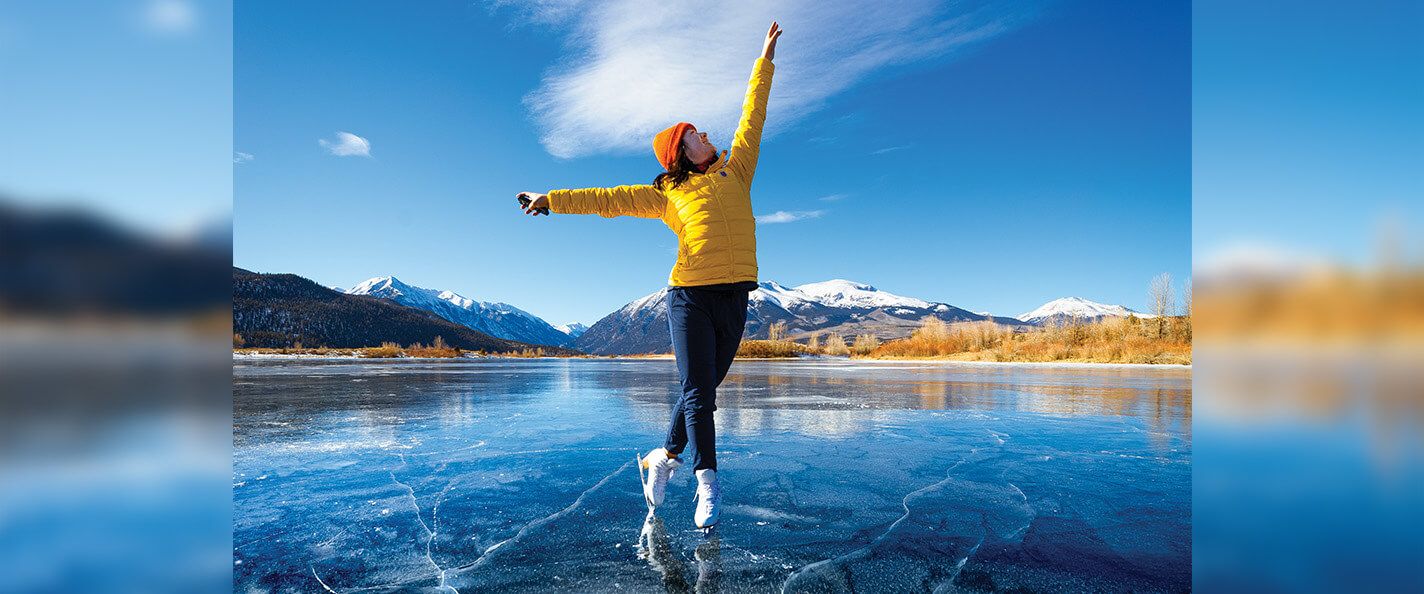 A woman on figure skates poses on a frozen lake. She's wearing an orange beanie, a yellow puffer coat, dark blue pants, and white figure skates. Her right leg is crossed just behind her left and balances on the toe. She is looking up and to the right, and her arms are extended up and to the right and out to the left. There are snow-capped mountains and blue sky on the horizon.