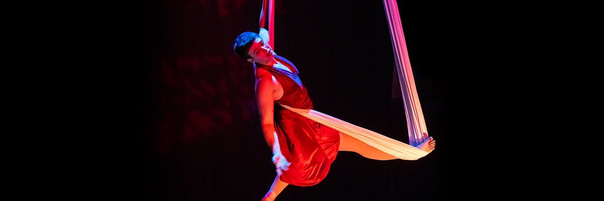A woman with short dark hair and wearing a red dress is suspended in a fabric sling. She is lit with a red stage light and set against a black background. Her left arm holds the sling, which wraps around her back and supports her outstretched left leg. Her right arm is stretched outward, and her right leg is straight under her.
