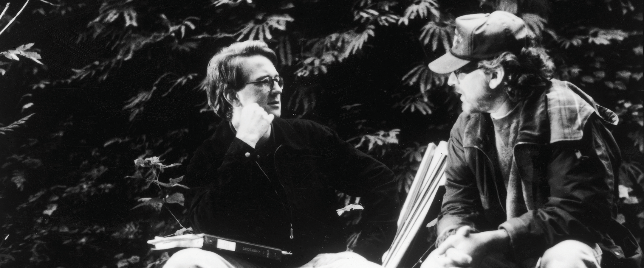 A black and white photo shows two men in casual dress sitting in front of a wall of greenery and facing each other in discussion.
