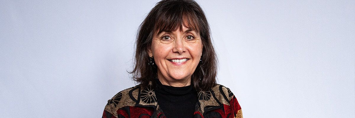 A woman is smiling against a white background. She has dark, shoulder-length hair and bangs. She wears a black shirt under a tan and red jacket with a black pattern and trim.