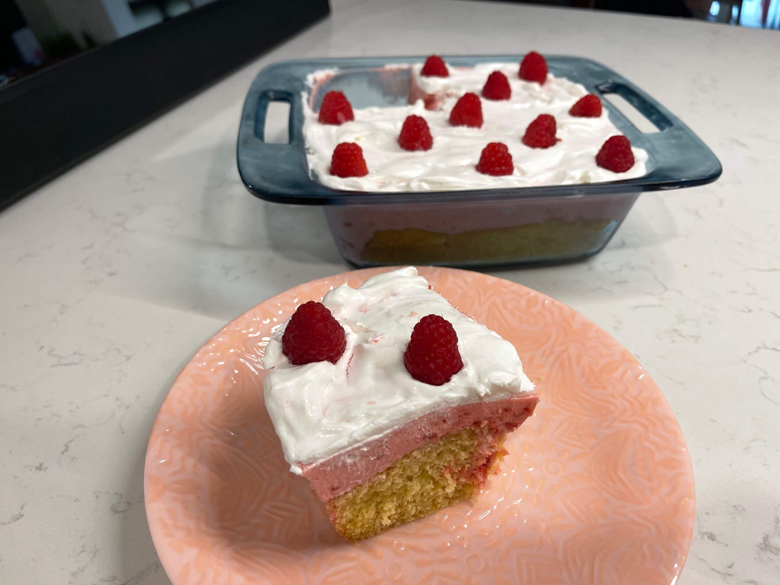 In the foreground, a square piece of yellow cake with white frosting and two raspberries on top sits on a round pink plate. In the background, a blue square dish contains the rest of the cake, iced white and dotted with raspberries. The back left corner piece is missing.