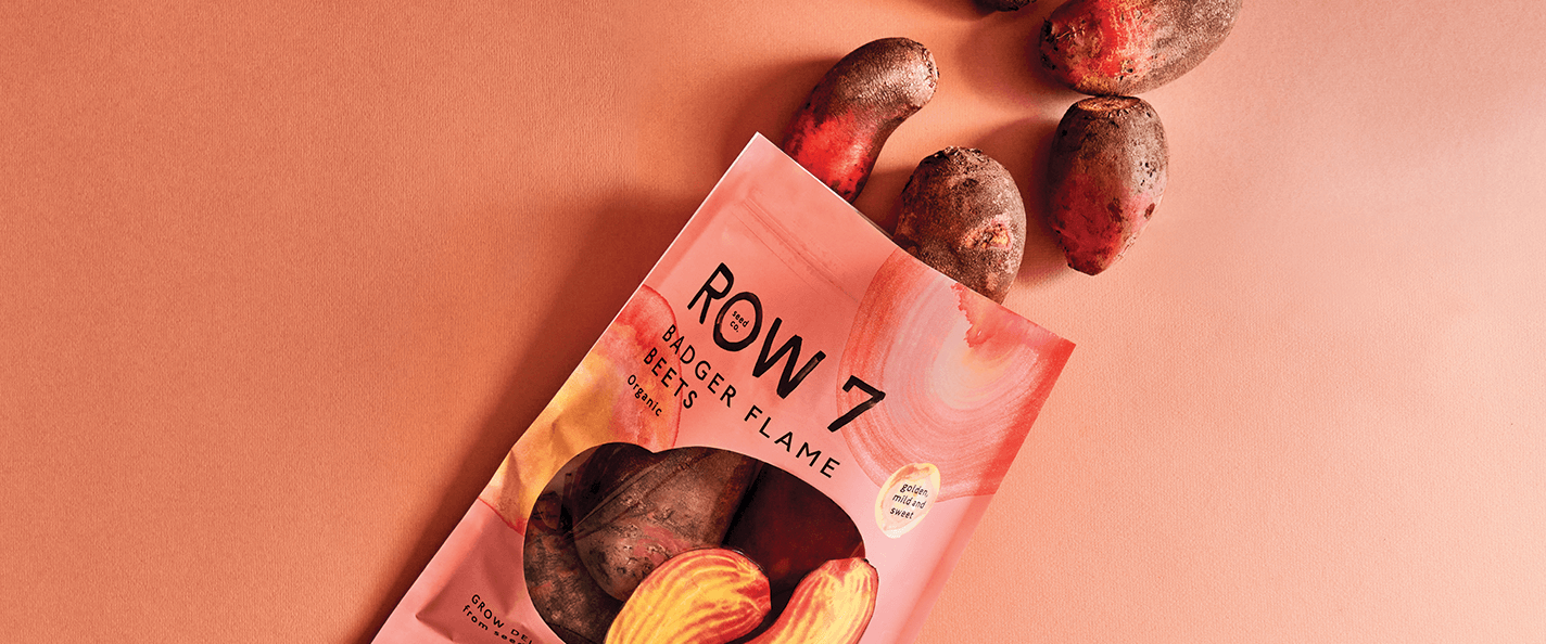 A coral-colored seed packet that says "Row 7" lies open on a coral-colored backdrop. There are beets visible through a transparent pane in the front of the packet and spilling out of the top of the packet.