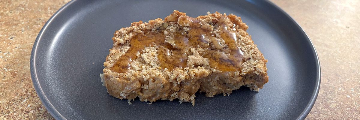 A light brown, textured loaf cake is drizzled with honey and sits on a dark grey, circular plate on a light wooden tabletop.