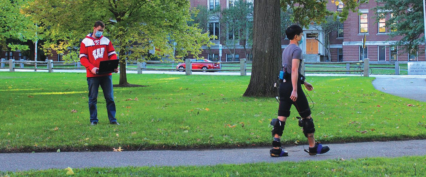 Two people are pictured on a university lawn. One individual is standing back and to the left looking at a laptop in his hands. He is wearing a red sweatshirt with a white W and jeans. The other individual is in the right foreground walking on the sidewalk. He is wearing a grey tshirt and black shorts. He is also wearing sensors on his torso, legs, and feet.