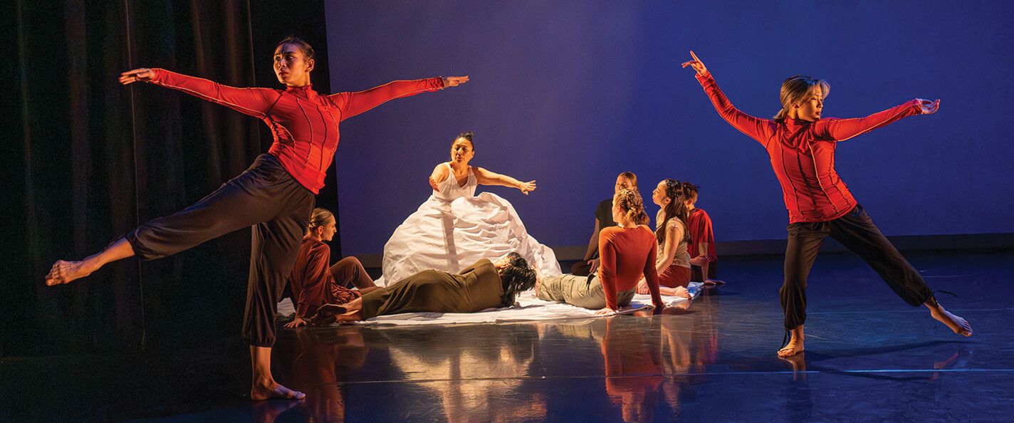 A dance performance on a stage. In the background, a woman in a white gown crouches and holds her arms out to dancers seated around her wearing plain, brown clothing. In the foreground, two dancers in fitted, red, long-sleeve tops and cropped black pants are positioned to the left and right of the woman in the gown and extend their arms and legs out and look off to the left and right, respectively.