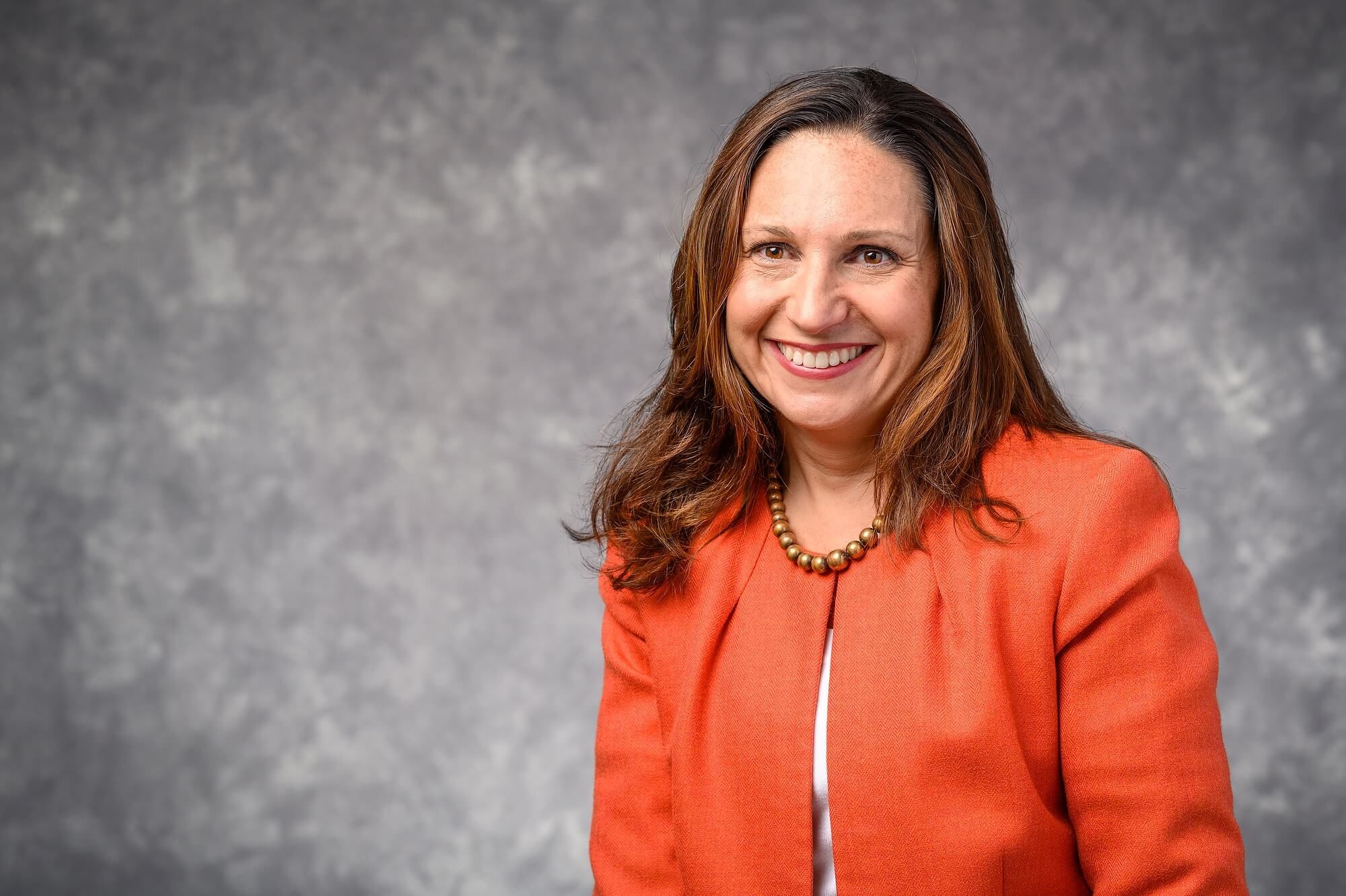 Christine Whelan is seated in the right side of the image against a mottled grey backdrop. She's wearing a red blazer and a chunky beeded necklace. Her brown hair is down around her shoulders.