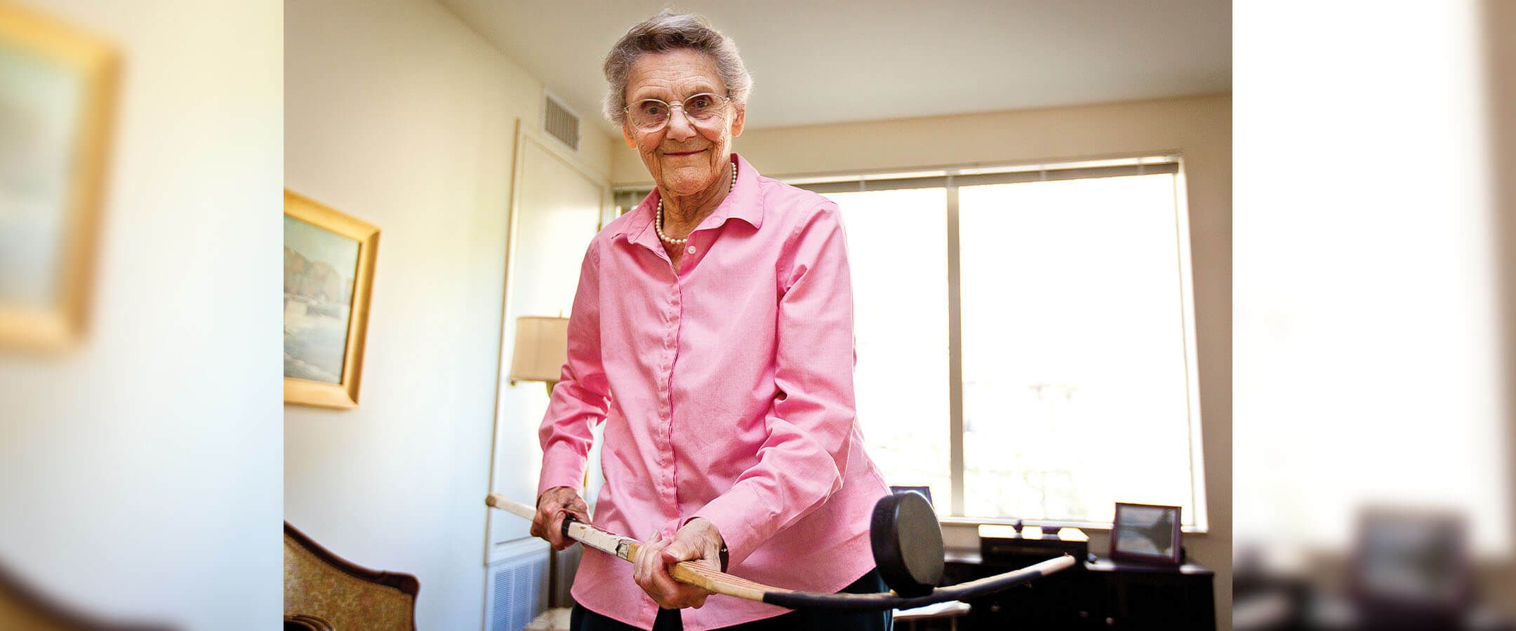 An elderly woman stands in an office holding a hockey stick on which she balances a hockey puck. She has short grey hair and round glasses, and she's wearing a pink button-up collared shirt. She is backlit by windows and looking/smiling down at the camera.