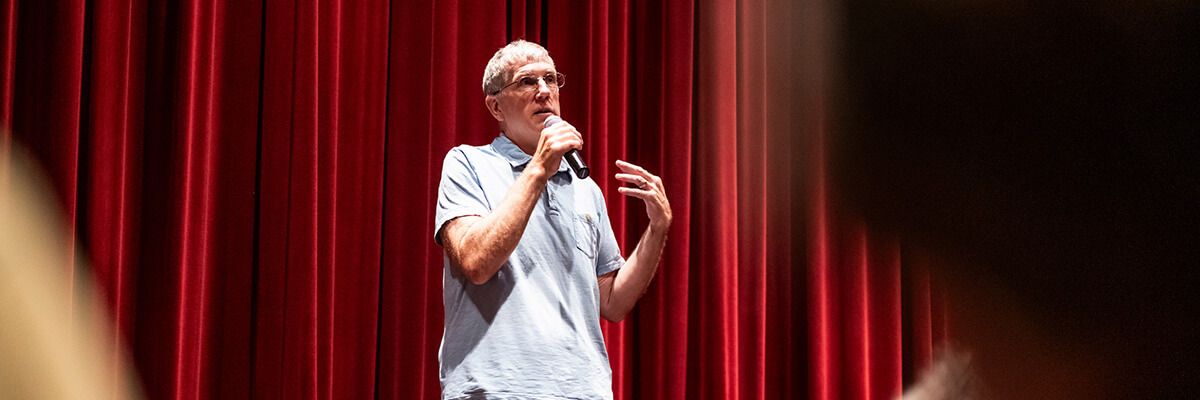 A man wearing a light-blue polo shirt and glasses is pictured standing in front of a red curtain and holding a microphone. He appears to be addressing an audience.