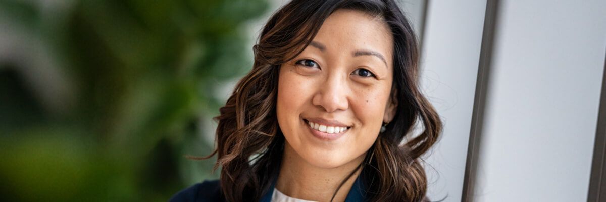 Kara is smiling, wearing a white blouse under a navy blue cardigan and light blue dangling stone earrings. Her shoulder-length hair is curled, and she's set against a blurred green backdrop with natural light coming in from the right.