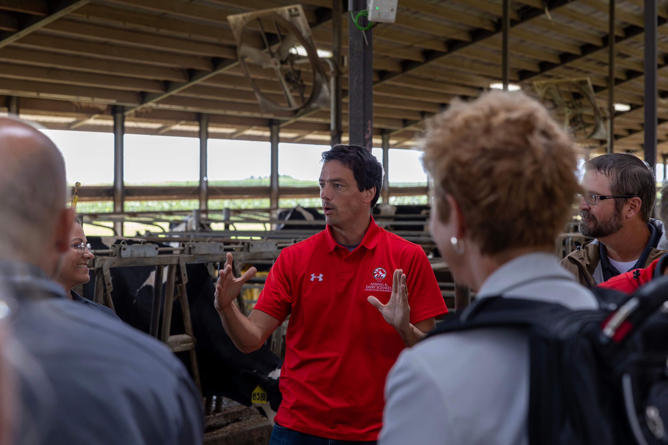 A man in a red polo stands in a high-ceilinged dairy barn and presents to a group of people who are standing in the foreground.