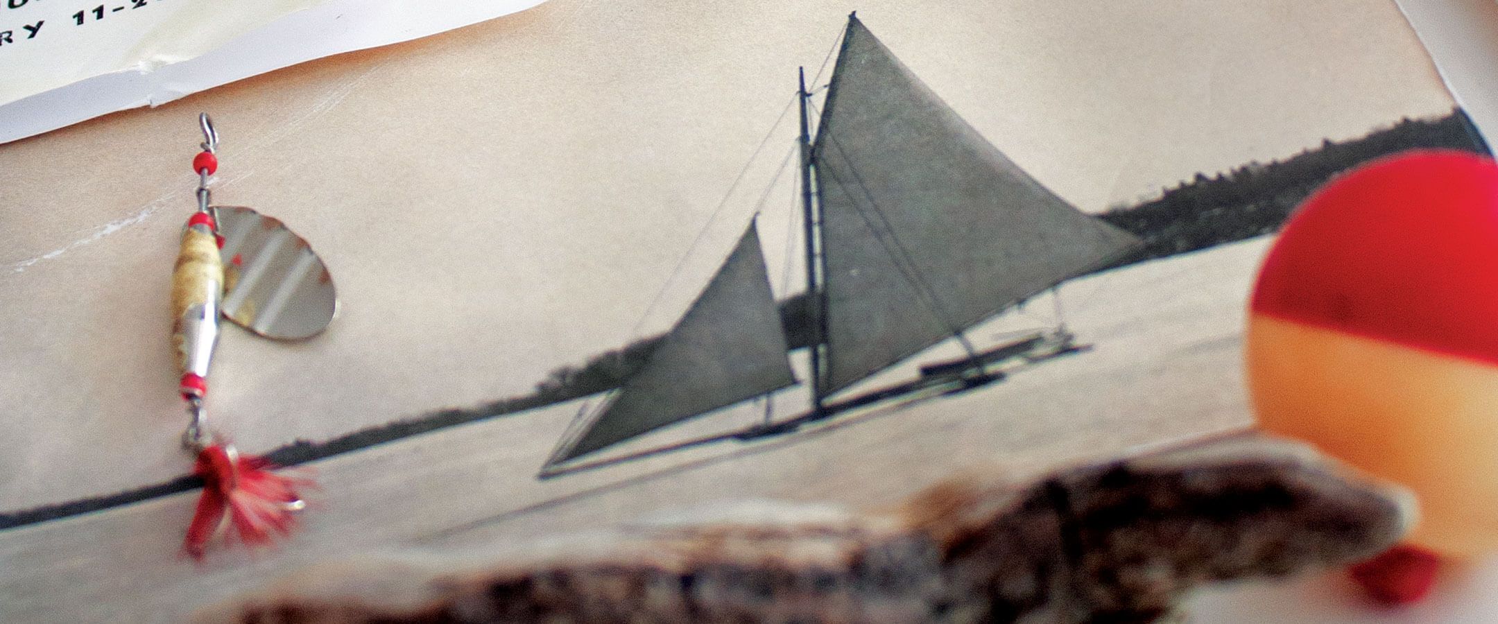 A fishing lure rests atop the left side of a photograph of a sailboat. There is a piece of driftwood and a bobber in the foreground in front of the photograph.