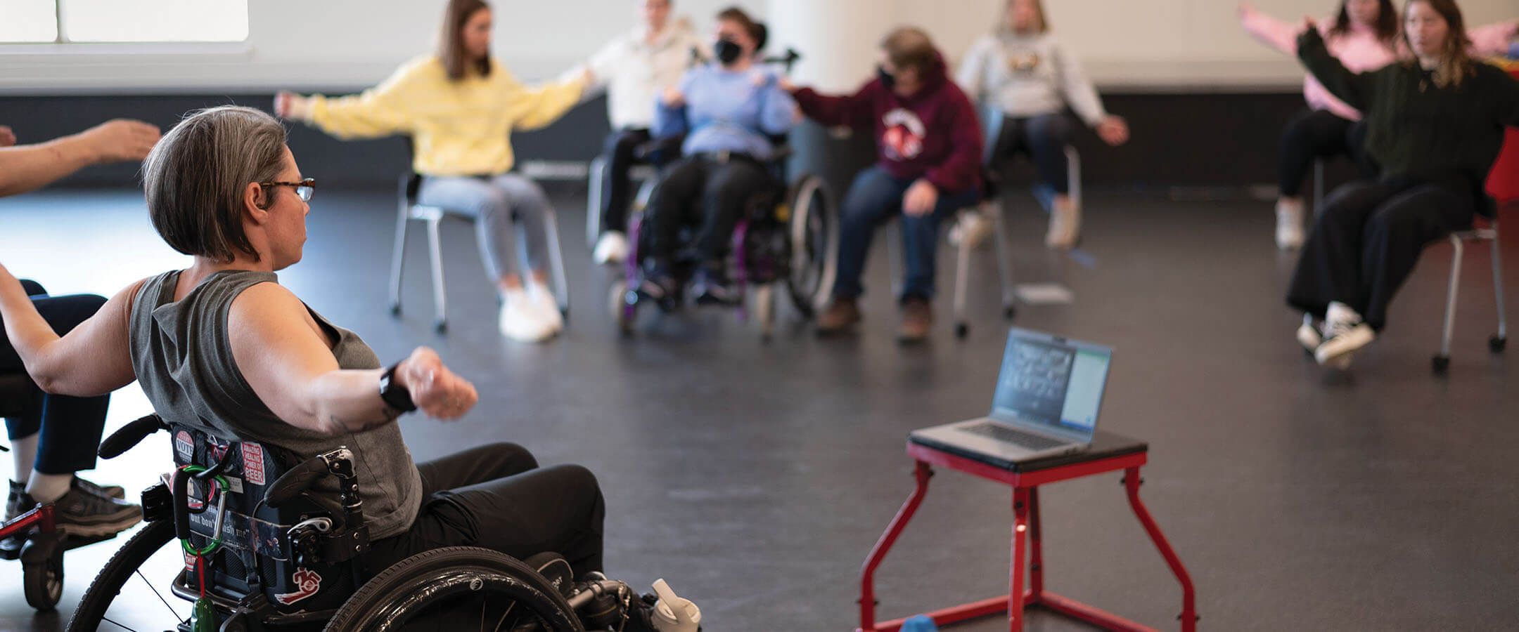 A woman leads a fitness class of seated individuals. The woman is pictured in the foreground in the bottom left of the image, sitting in a wheelchair and facing away from the camera. She has short, dark hair and glasses, and she is wearing a grey sleeveless shirt and black pants. Her arms are outstretched to either side. In front of her, a laptop sits open on a red stool, broadcasting her instruction to viewers online. Behind the laptop, a group of seated individuals (out of focus) mirror the instructor's movements.
