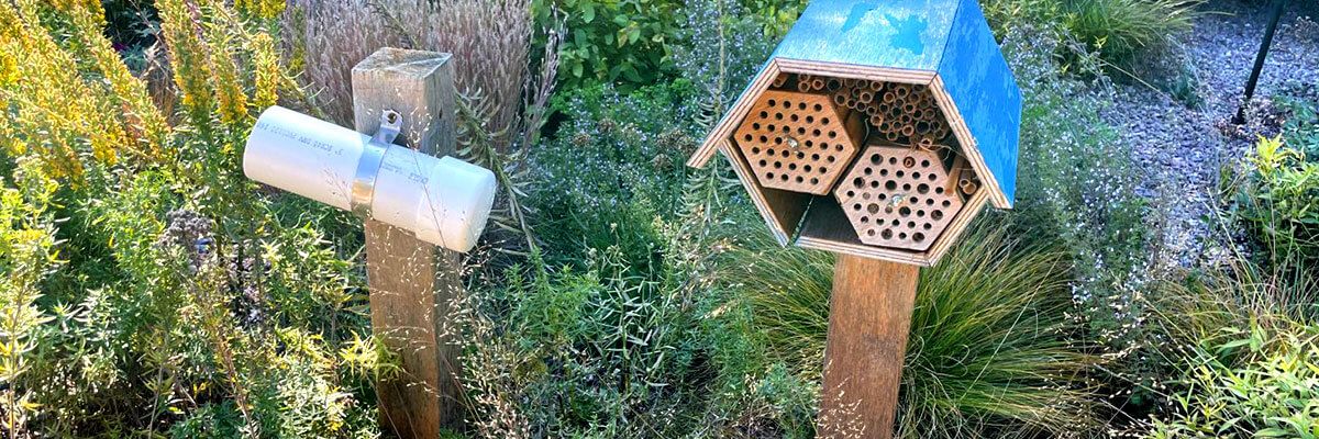 Two wooden posts are set side-by-side in the ground in a garden space, surrounded by tall, green grasses. The sun is shining in from the left side of the frame. On the post to the left is mounted a short length of white tubing. On the right post is mounted a hexagonal container with an open face. Inside are two smaller hexagons, staggered. Each smaller hexagon has a front cover with several small holes. In the space above the smaller hexagons, there are reeds/wooden tubes stacked to the top of the larger hexagon.